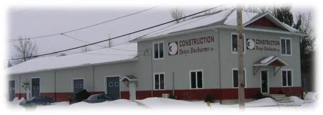 Bâtiment blanc à deux étages portant l’enseigne « Construction Denys Ducharme », en hiver.